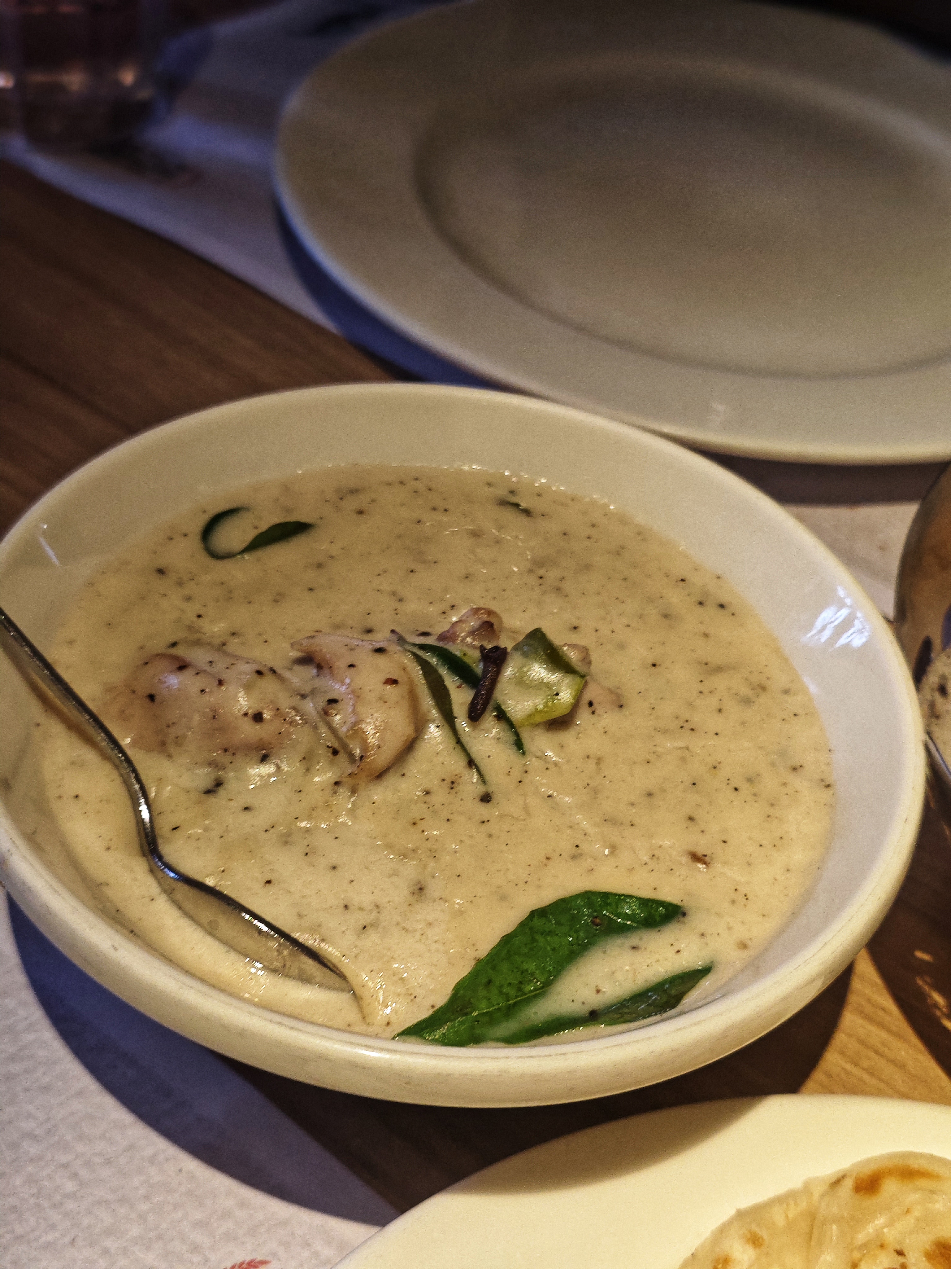 A white bowl of creamy, beige soup with herbs and chicken pieces. A spoon rests inside.