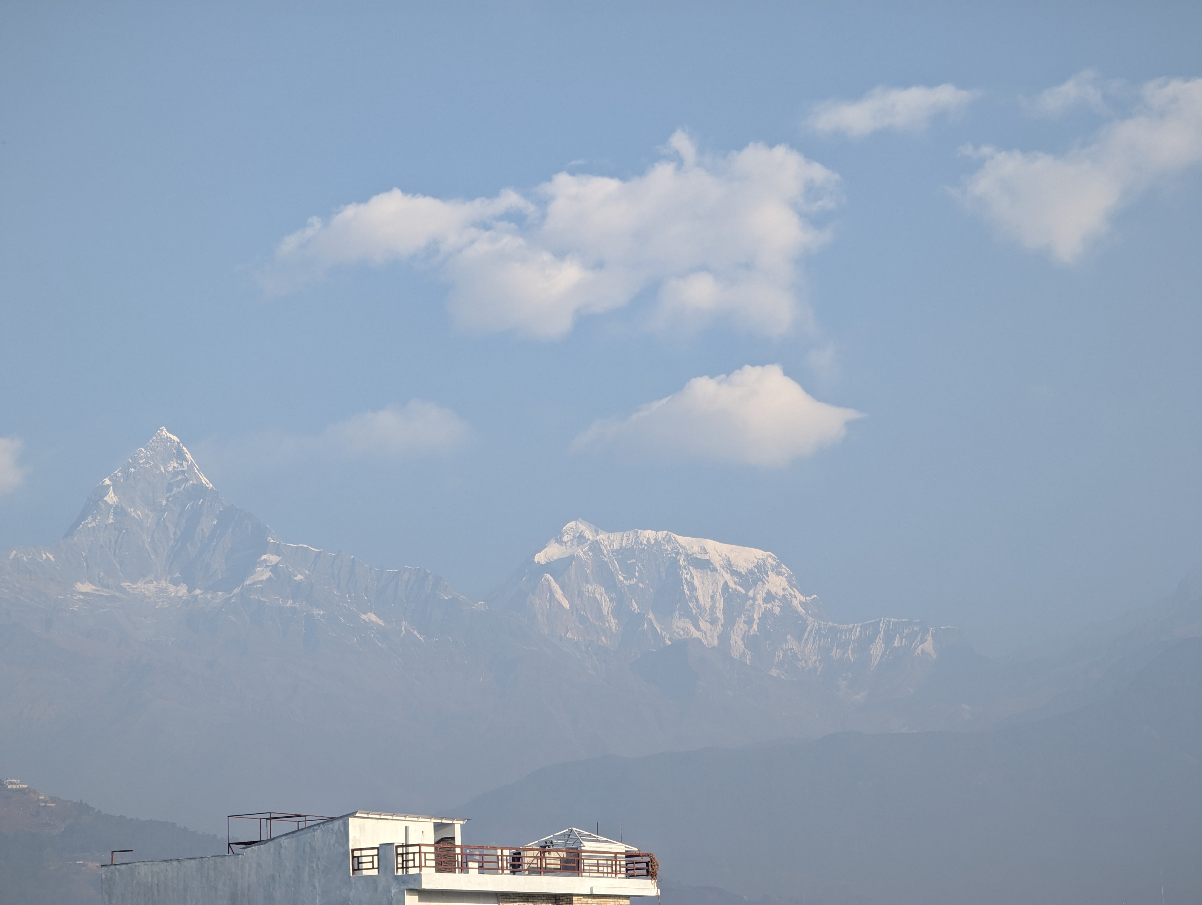 A majestic, snow-capped pyramid peak and rugged mountains rise against a light blue sky with wisps of clouds, viewed from a rooftop with a minimal structure.