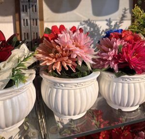 Vibrant artificial flowers in shades of pink and red fill ornate white pots on a glass shelf. Sunlight casts soft shadows, creating a warm, inviting ambiance.