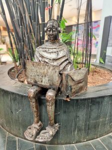 A metal sculpture of a seated Masai lady figure holding a laptop and bag beside tall bamboo stems.