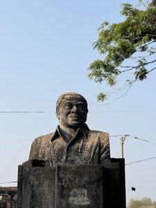 A bronze calicut statue on a pedestal, surrounded by greenery under a clear blue sky.
