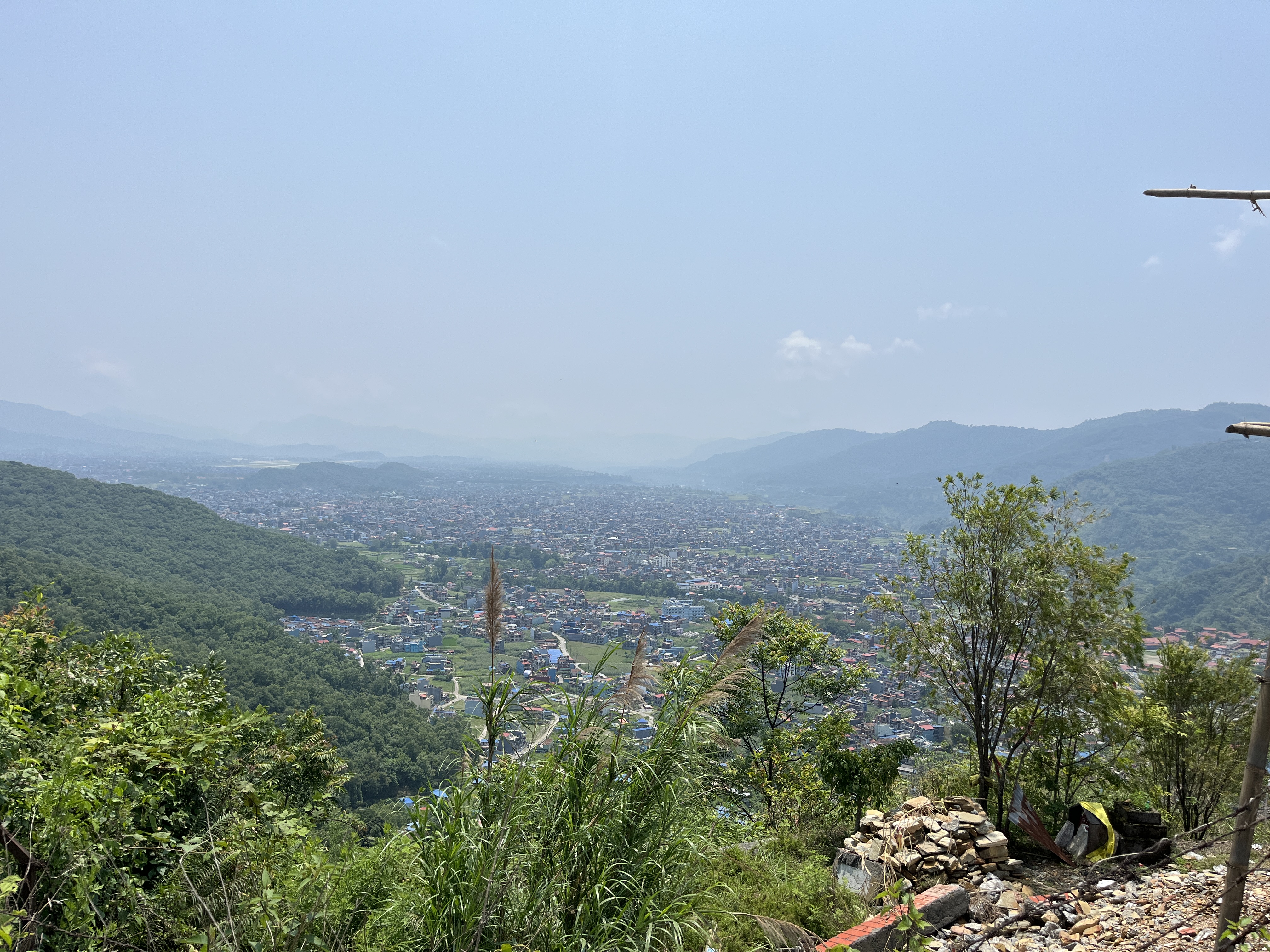 View of a Pokhara city from a hillside with lush green trees and grass in the foreground, mountains in the background, under a bright, clear sky.