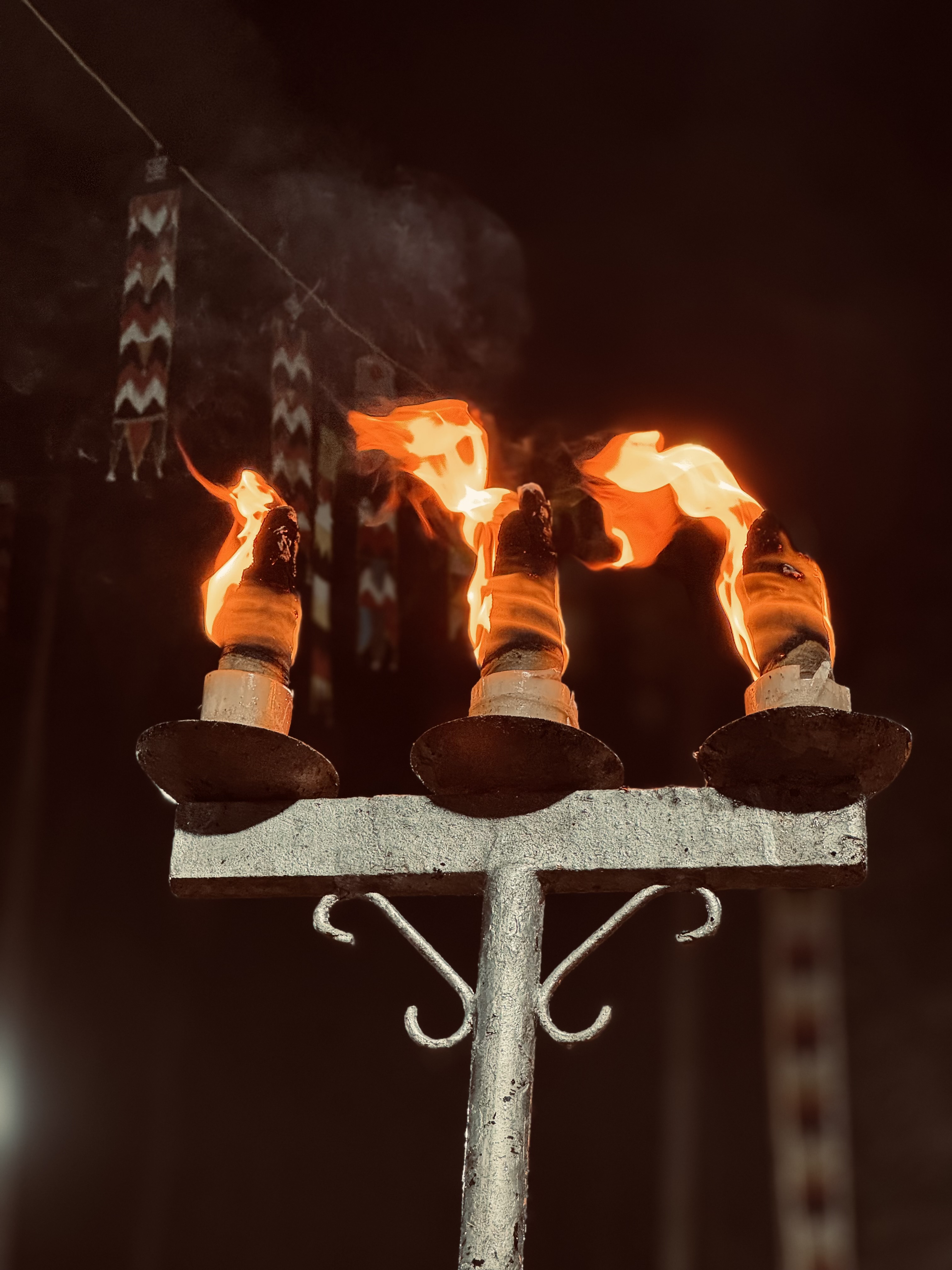 Three flaming vilakku torches are mounted on a metallic stand, casting a warm glow against a dark background. The flames are vibrant and flickering, with wisps of smoke rising into the night air.