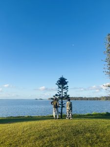 An image of two people standing and looking toward the beautiful view of the sea. Behind them is a green garssy land.
