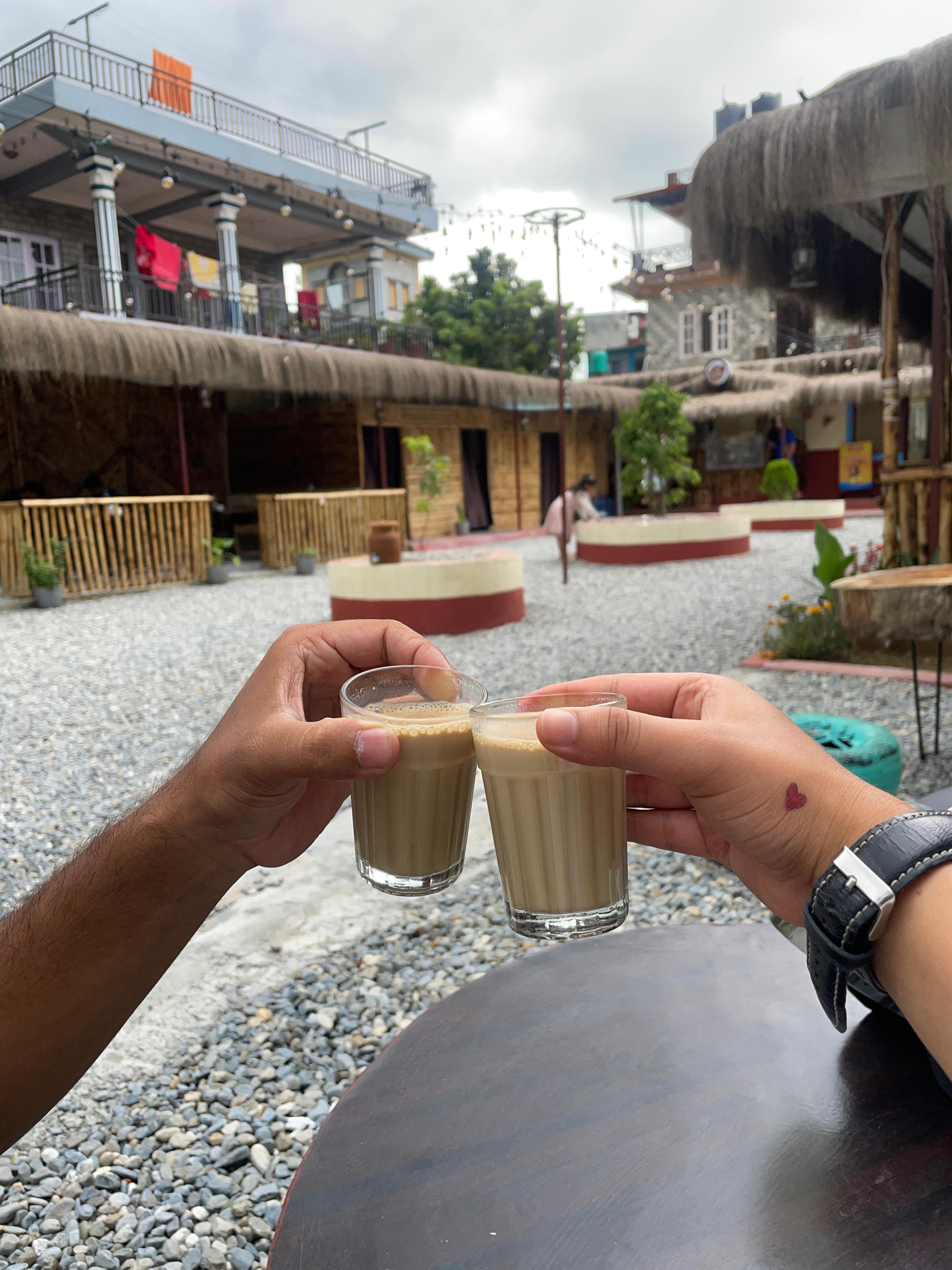 A close-up of two people's hands gently clinking clear glasses of milk tea over a dark table. The person on the right has a small red heart tattoo on their wrist and wears a black watch. In the background, a gravel courtyard features bamboo structures with thatched roofs under a cloudy sky.