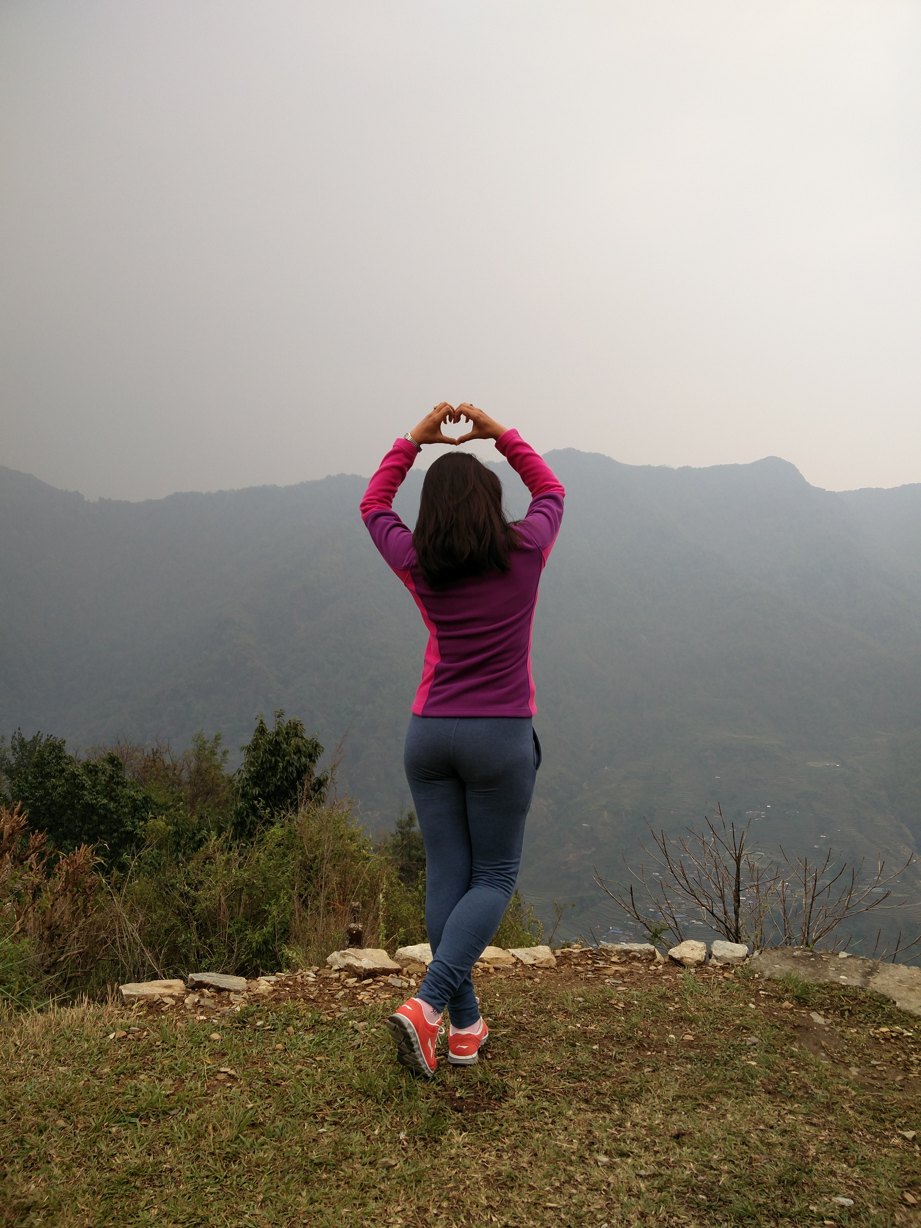 A woman with her back to the camera stands on a grassy overlook, making a heart shape with her hands against a backdrop of misty mountains.