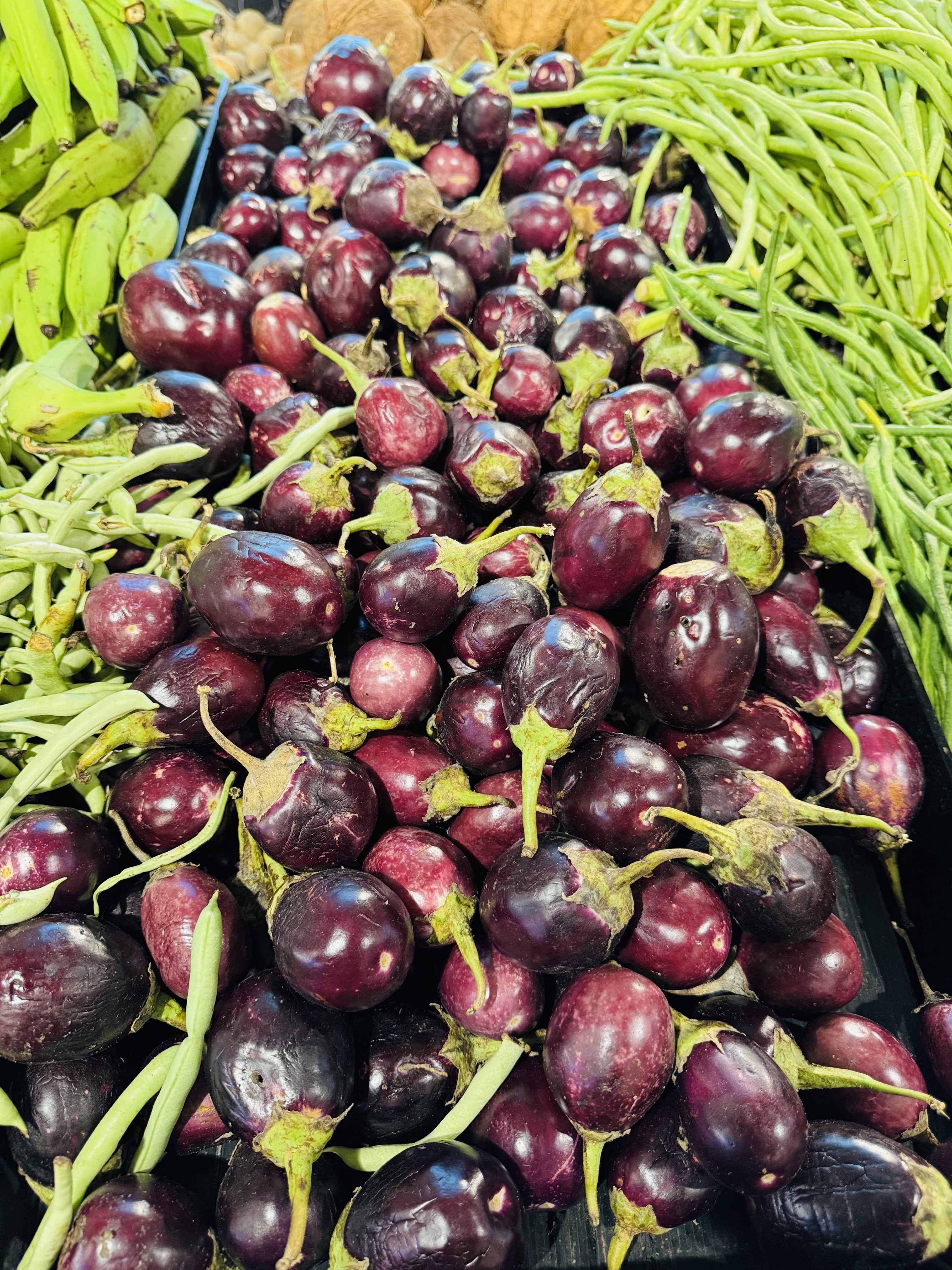 A vibrant pile of small purple eggplants is surrounded by green beans and bananas.