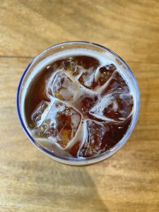 A glass of iced coffee with bubbles on the surface sits on a wooden table.