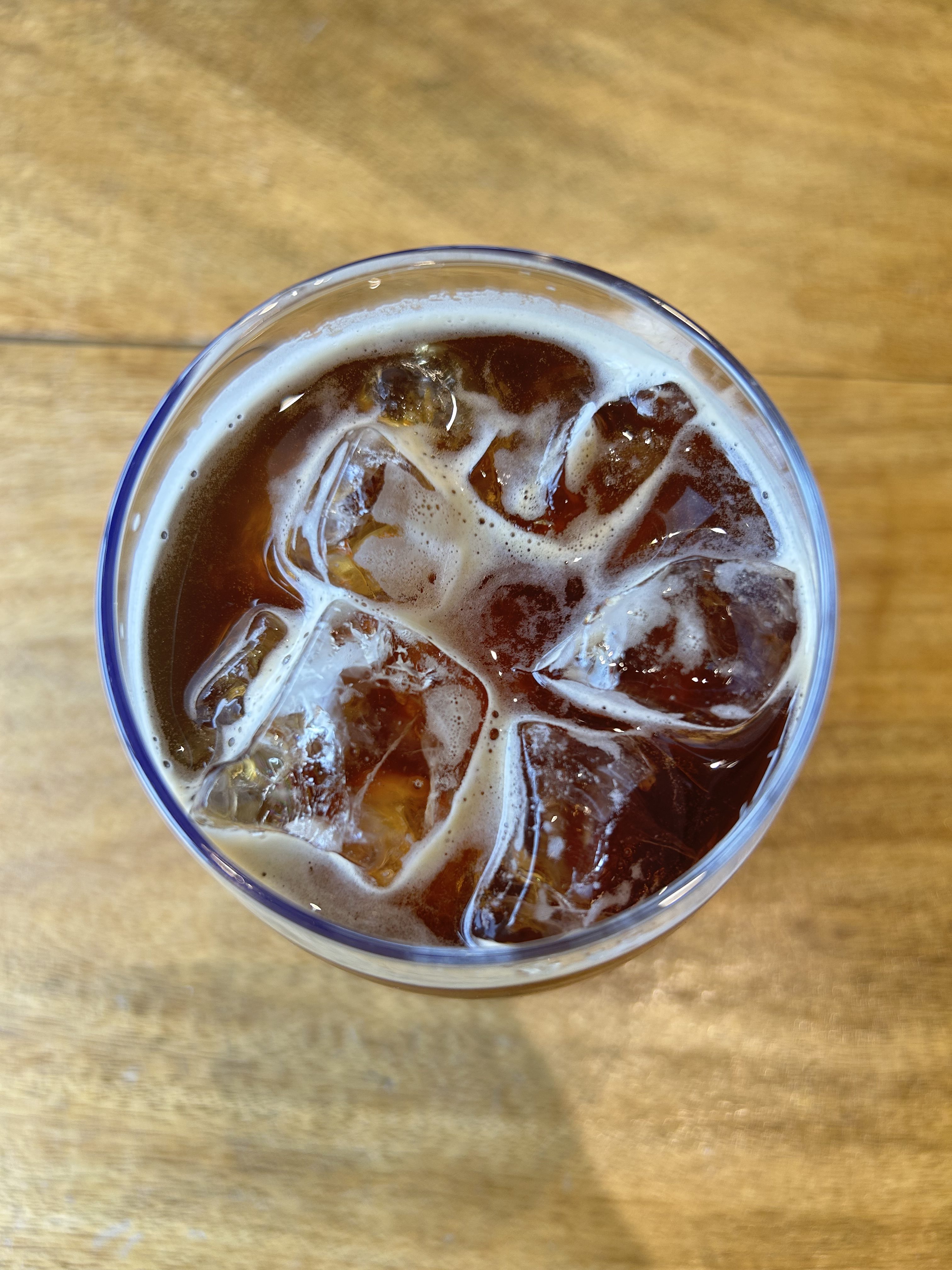 A glass of iced coffee with bubbles on the surface sits on a wooden table.