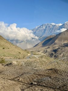 Mountain landscape with a winding road, arid terrain, and a rocky hillside. Snow-capped peaks and fluffy clouds are set against a bright blue sky.