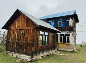 A rustic weathered two-story wooden house with a blue metal roof stands on grassy ground, with prayer flags adding a serene cultural touch.