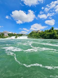 A wide landscape view shows the turquoise Rhine River, white spray from a waterfall, green hills, and a blue sky with soft clouds. Boats and historic buildings complete this stunning scene near the Rhine Falls, Switzerland.