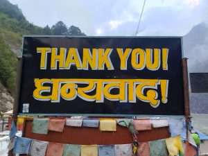 A bold “Thank You” sign in yellow letters with colorful prayer flags hanging below, set against a mountain backdrop.