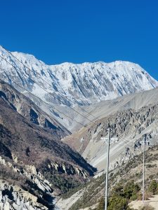 Snow-capped mountains under a clear blue sky, with rugged terrain and sparse vegetation in the foreground. Power lines stretch across the scene.