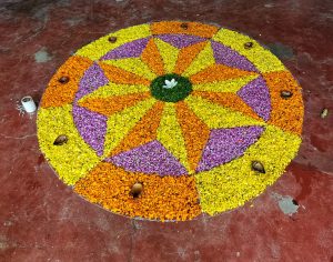 Colorful flower rangoli with yellows, oranges, and purples forms a geometric pattern on a red floor, centered by a white flower, creating a festive, vibrant atmosphere.
