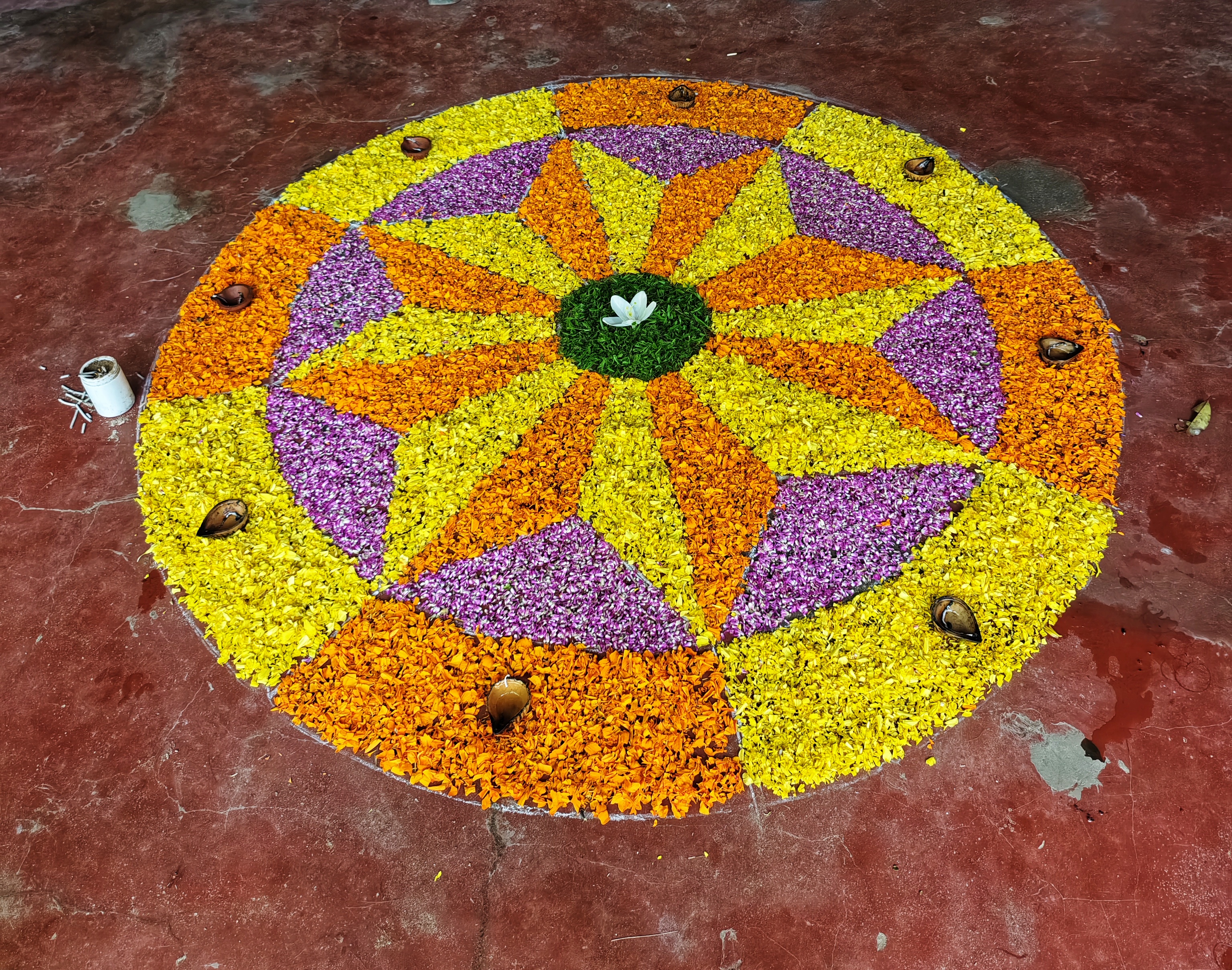 Colorful flower rangoli with yellows, oranges, and purples forms a geometric pattern on a red floor, centered by a white flower, creating a festive, vibrant atmosphere.