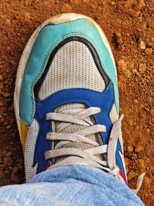 A close-up view of a colorful sneaker with a blue, green, and yellow design, resting on a patch of reddish-brown dirt. 