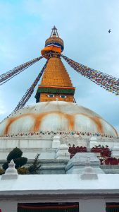 Low-angle view of Boudhanath Stupa with Buddha eyes, colorful prayer flags, and a white dome under a cloudy blue sky.
