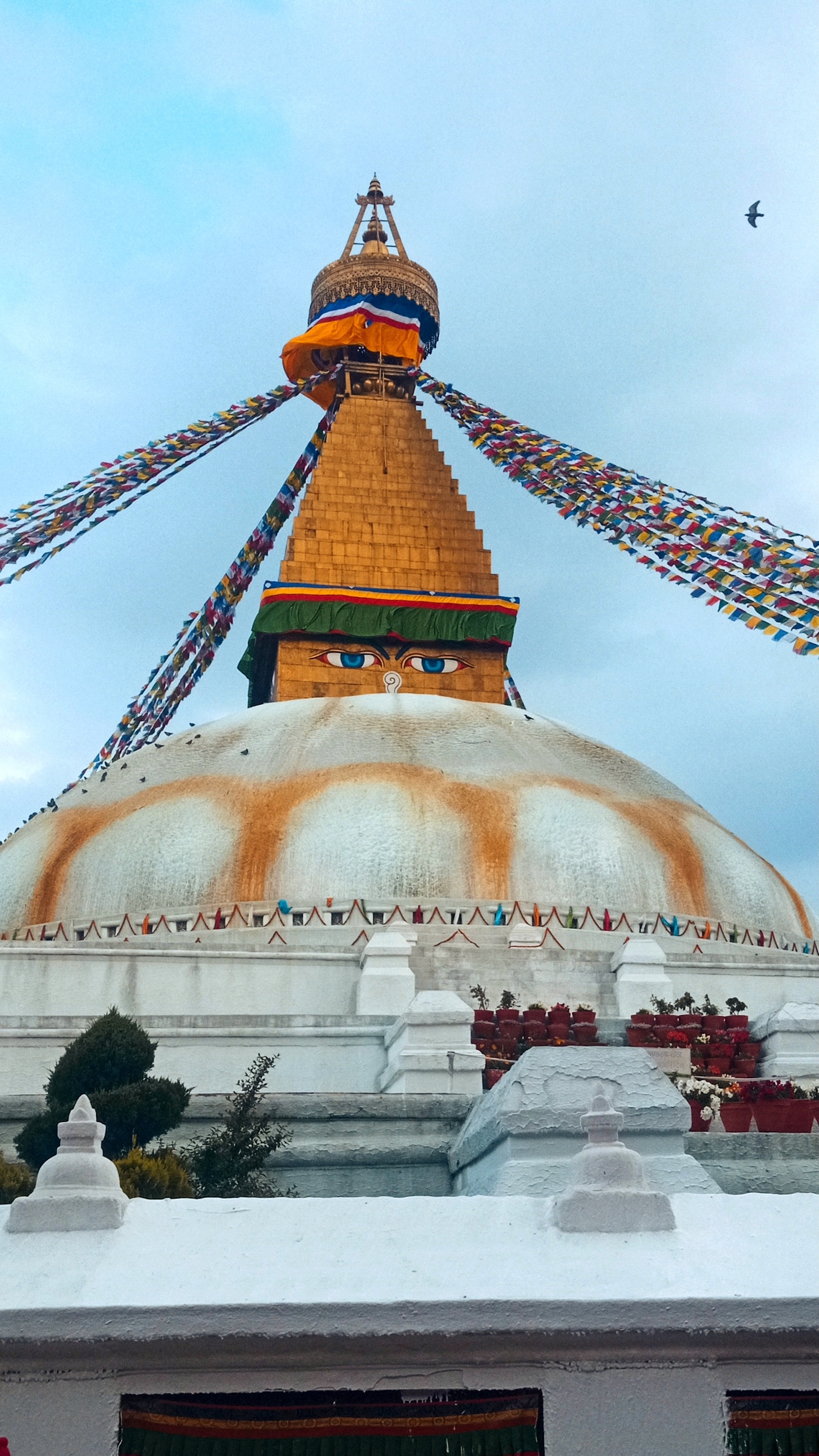 Low-angle view of Boudhanath Stupa with Buddha eyes, colorful prayer flags, and a white dome under a cloudy blue sky.