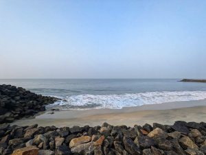 A tranquil beach scene featuring a calm ocean under a pale blue sky. In the foreground, clusters of large, dark rocks line the shore, while gentle waves wash onto the sandy beach. 