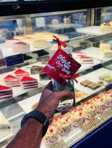 A hand holding a glass bottle with a red tag reading "My one &amp; only," in front of a pastry display case filled with colorful cakes and desserts.