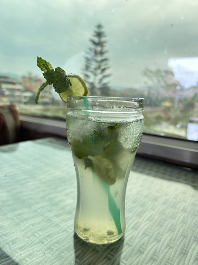 A refreshing mojito in a curvy glass with ice, mint leaves, and a lime slice garnish. A green straw is submerged in the drink, placed on a table by a window.