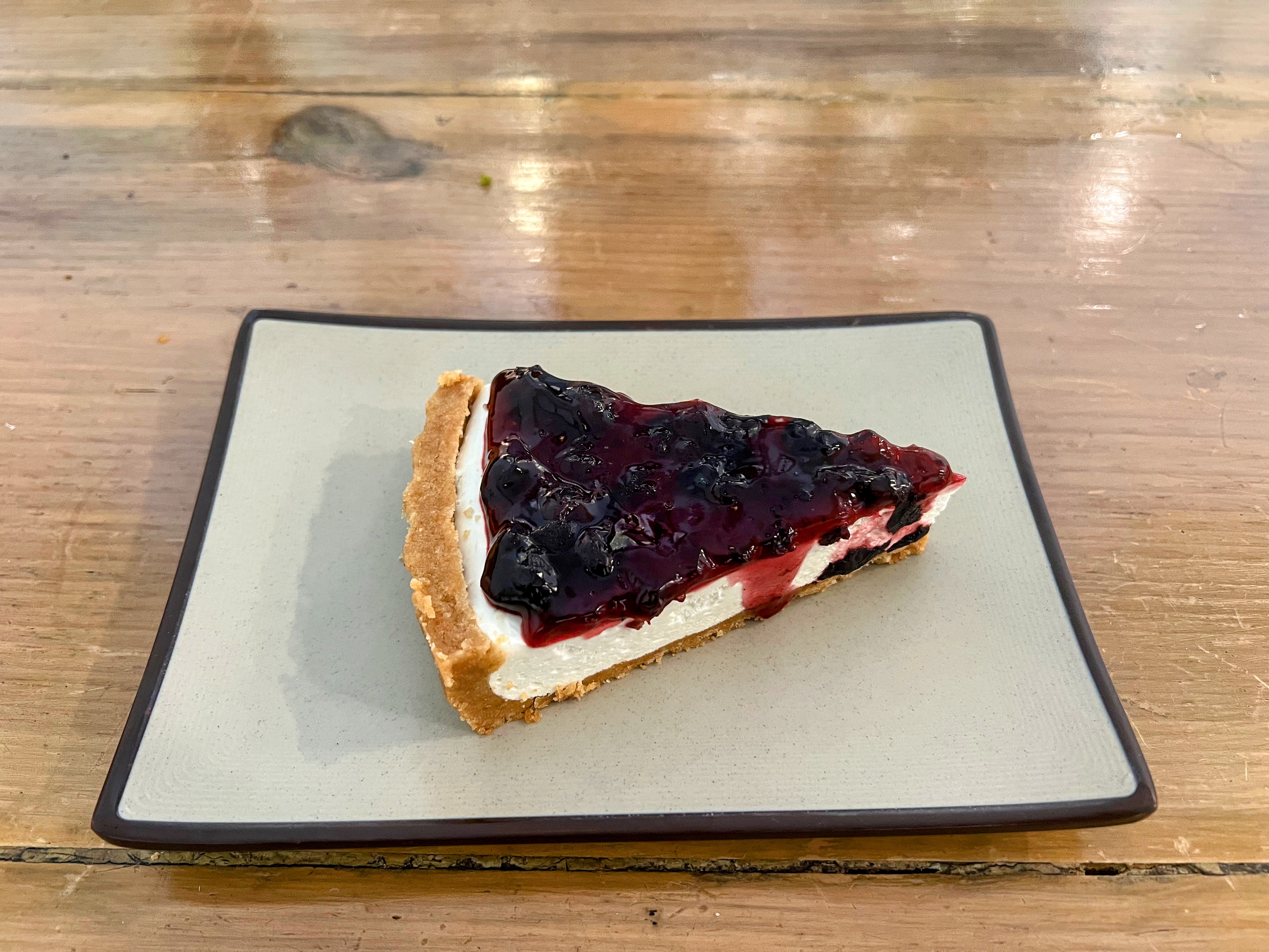 A slice of cheesecake topped with a thick layer of glossy blueberry compote, served on a square, light-gray plate over a wooden table.