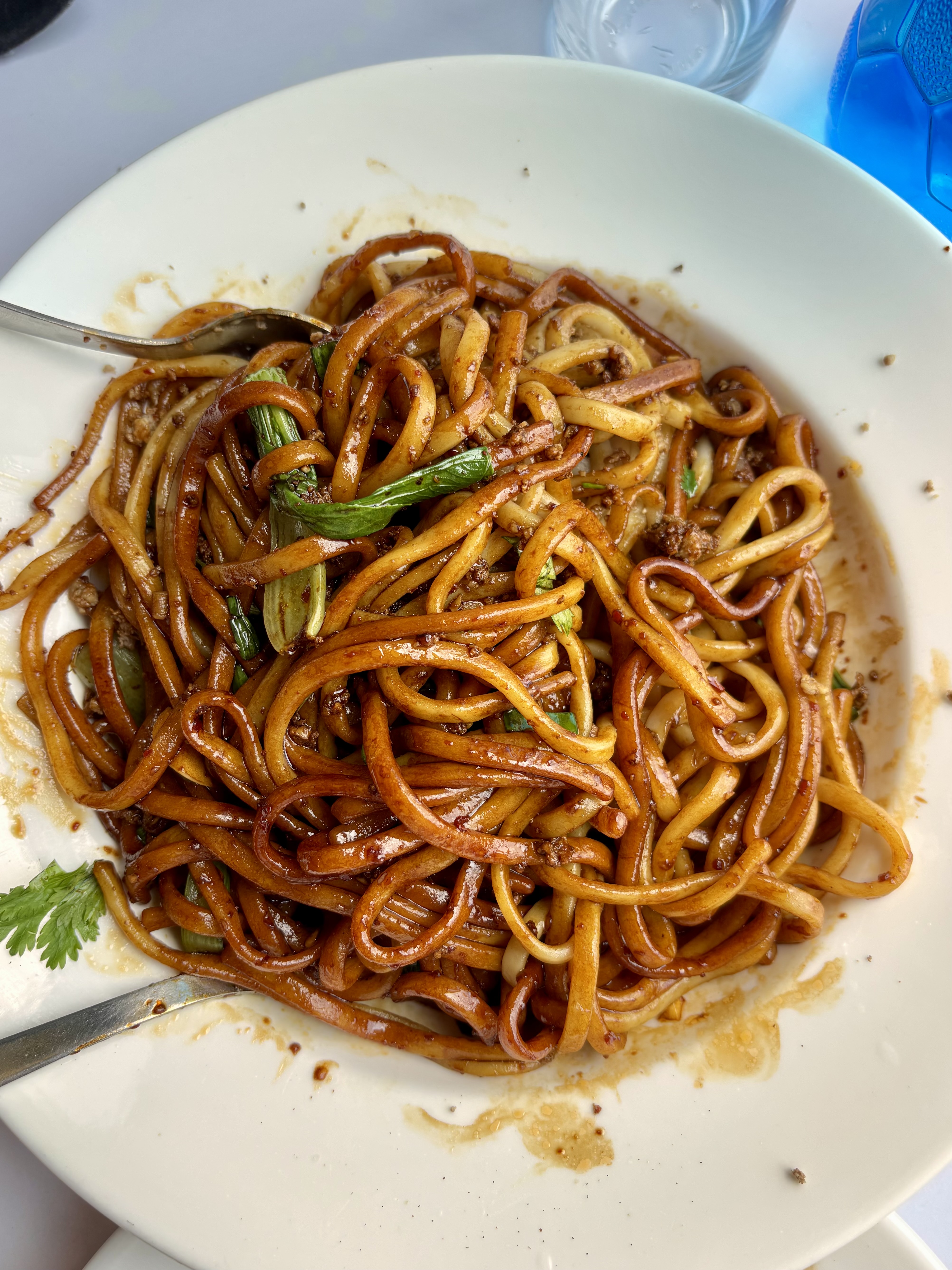 A plate of stir-fried noodles with green vegetables and sauce, served with a fork and spoon.