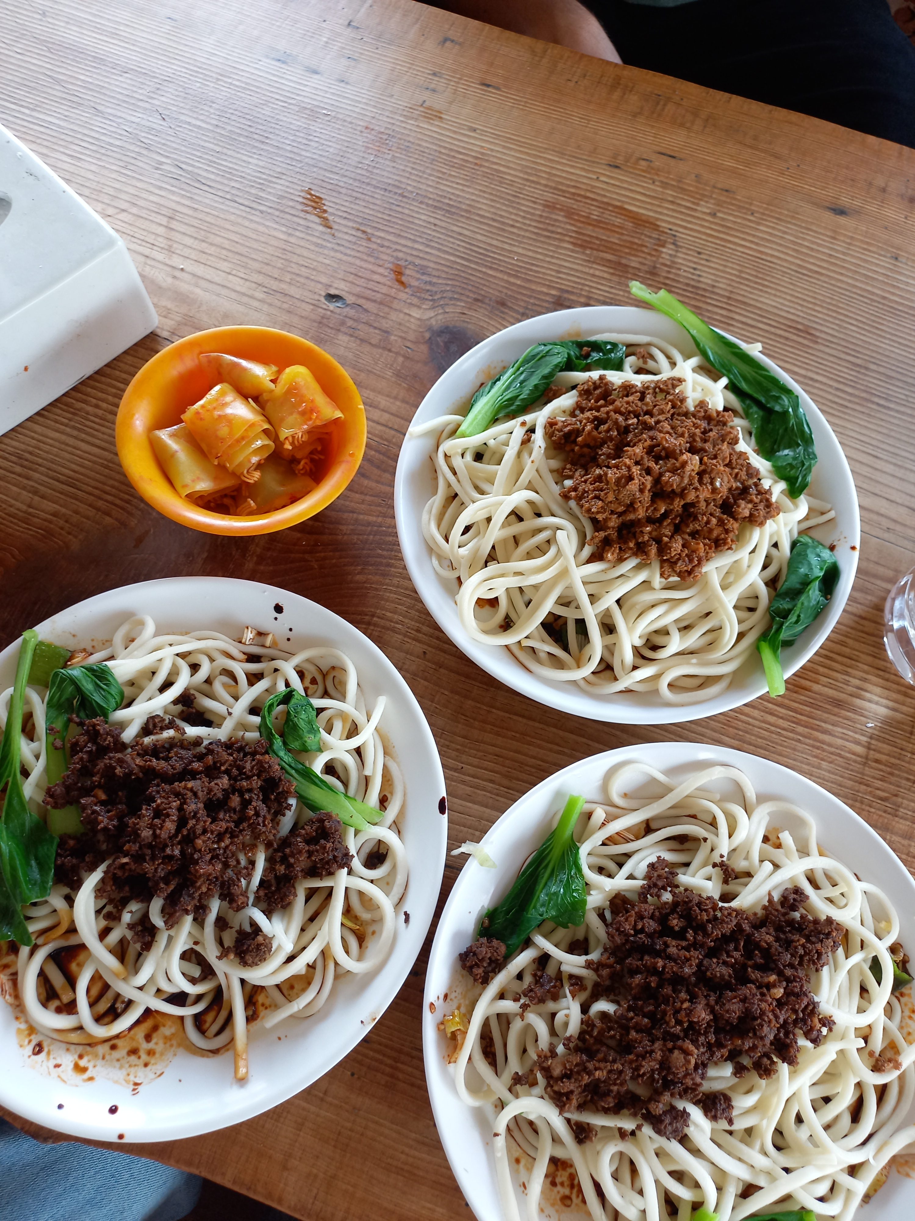 A top-down shot of a wooden table featuring three white bowls filled with thick noodles, each topped with a dark minced meat sauce and fresh green bok choy. A small orange bowl containing a spicy side dish sits near a white tissue box on the left.