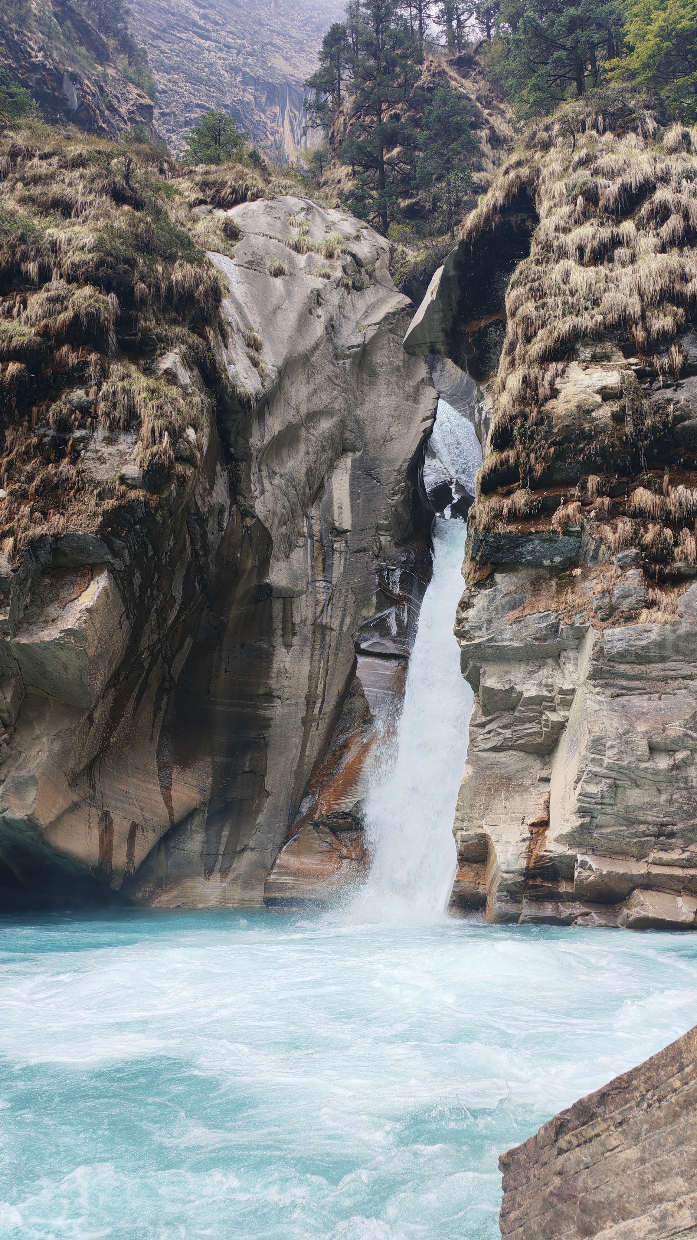 A powerful waterfall cascades through a narrow rock formation, surrounded by rugged cliffs covered in moss, flowing into a vibrant turquoise river.