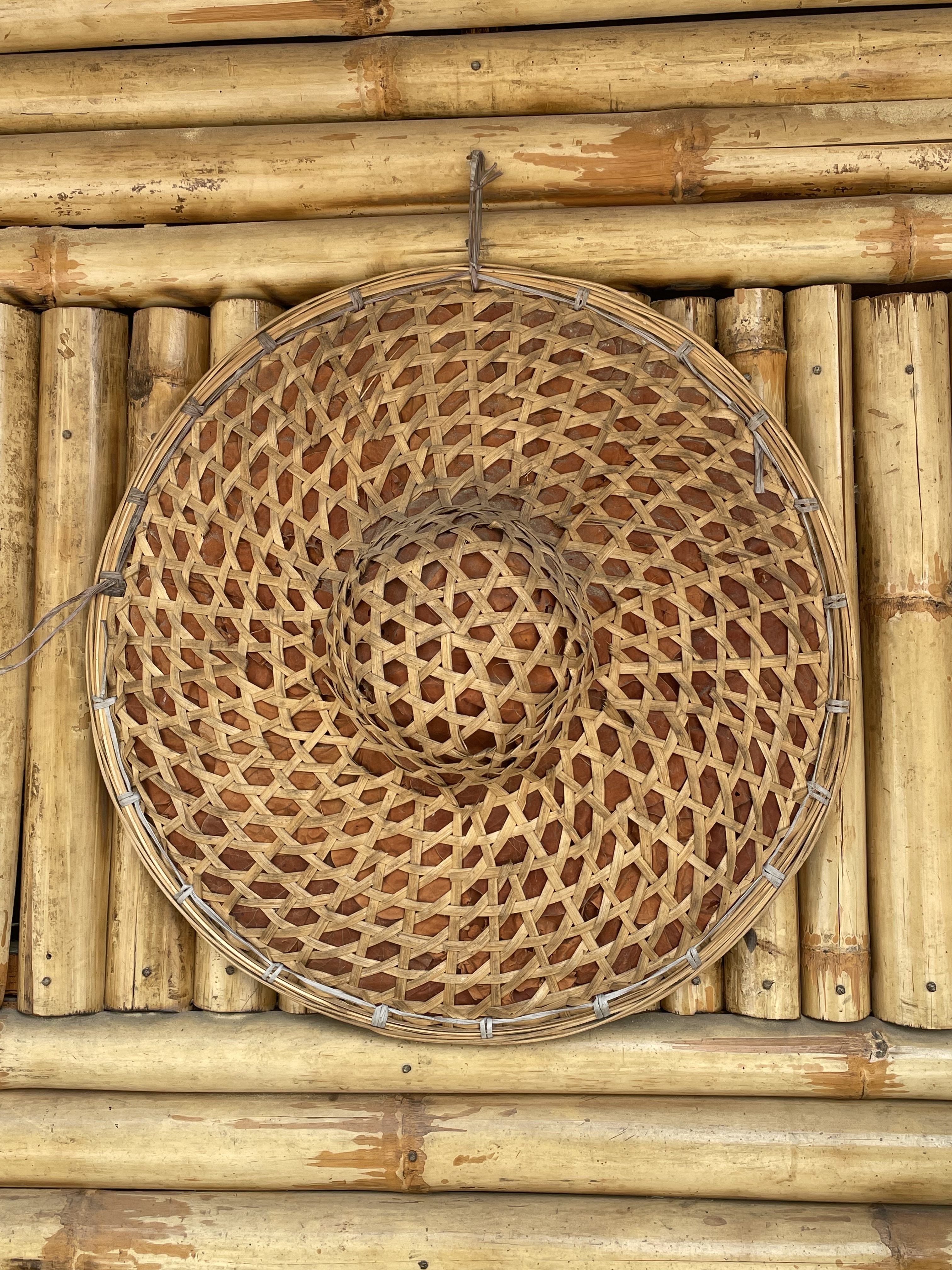 A front-facing close-up of a traditional, circular woven bamboo hat with a raised center crown, hanging on a wall made of horizontal bamboo stalks.