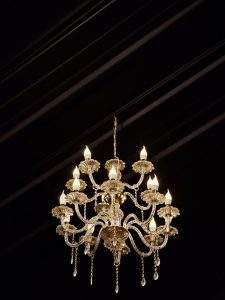A beautiful silver-colored chandelier hangs from a dark ceiling, glowing warmly and adding a rich, elegant feel to the space. Shot indoors in Perumanna, Kozhikode. 