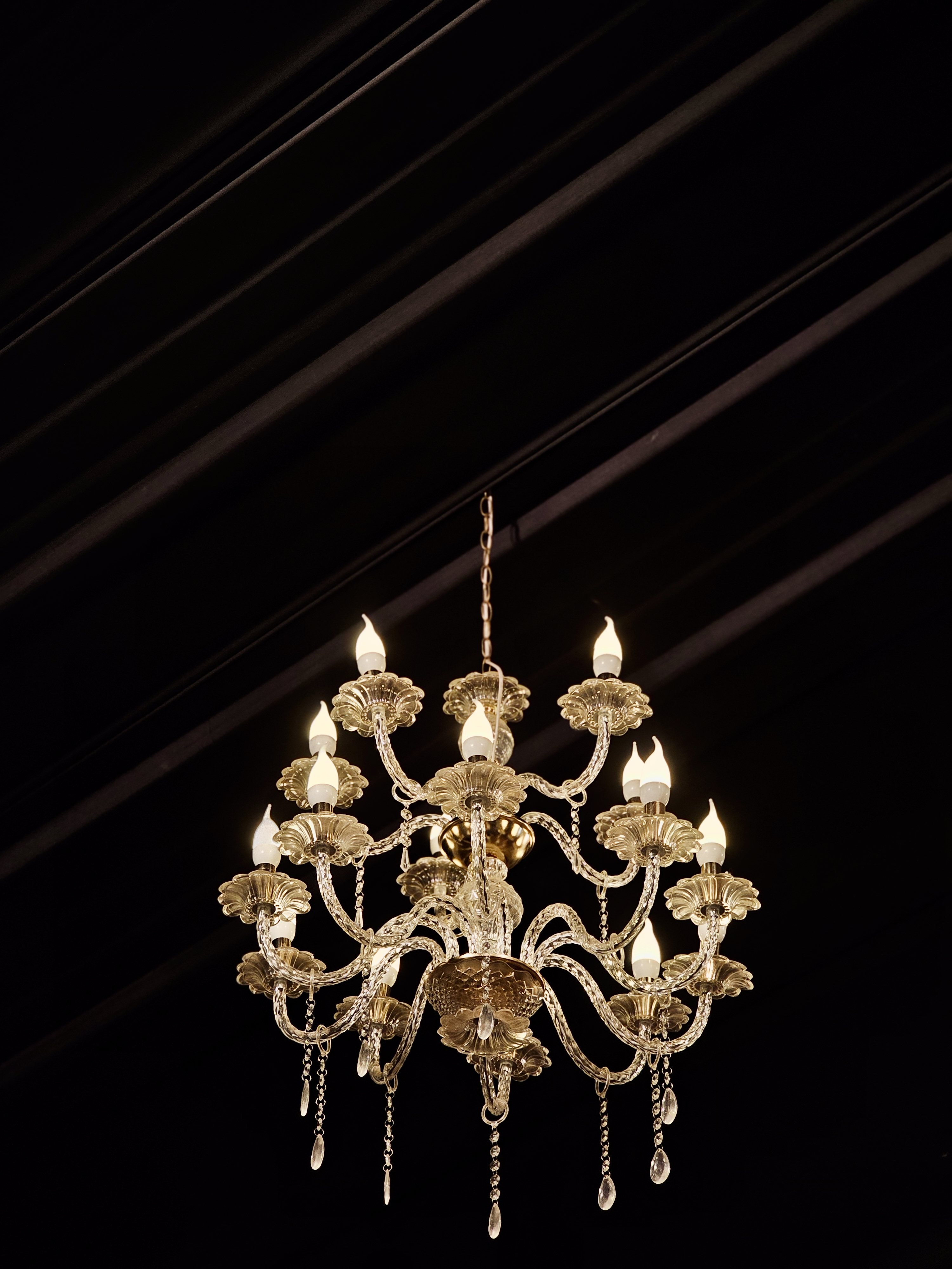 A beautiful silver-colored chandelier hangs from a dark ceiling, glowing warmly and adding a rich, elegant feel to the space. Shot indoors in Perumanna, Kozhikode.