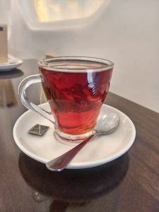 A clear glass cup of tea on a white saucer with a spoon and tea bag.