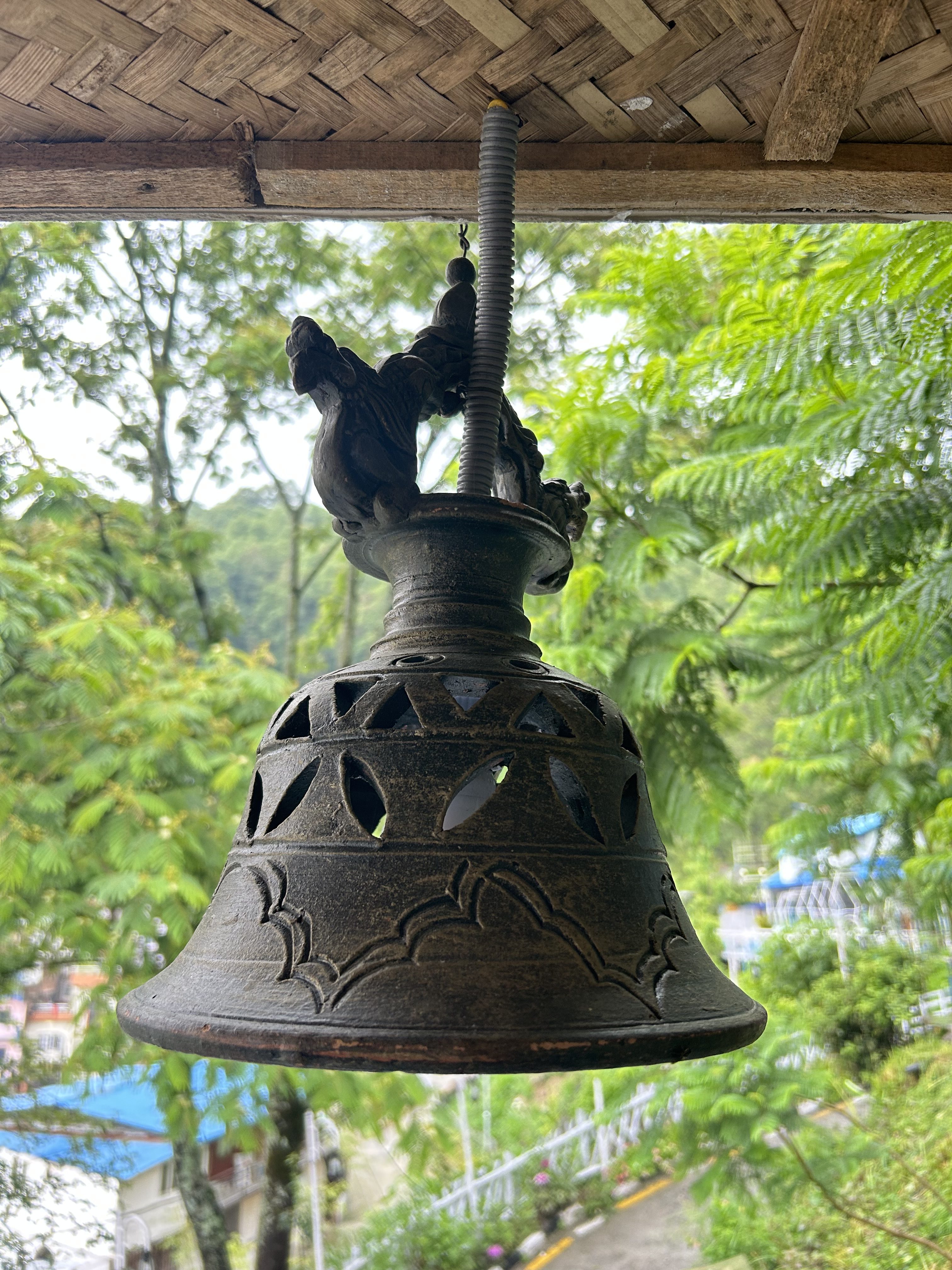 Ornate bronze bell hanging from a wooden ceiling, surrounded by lush greenery.