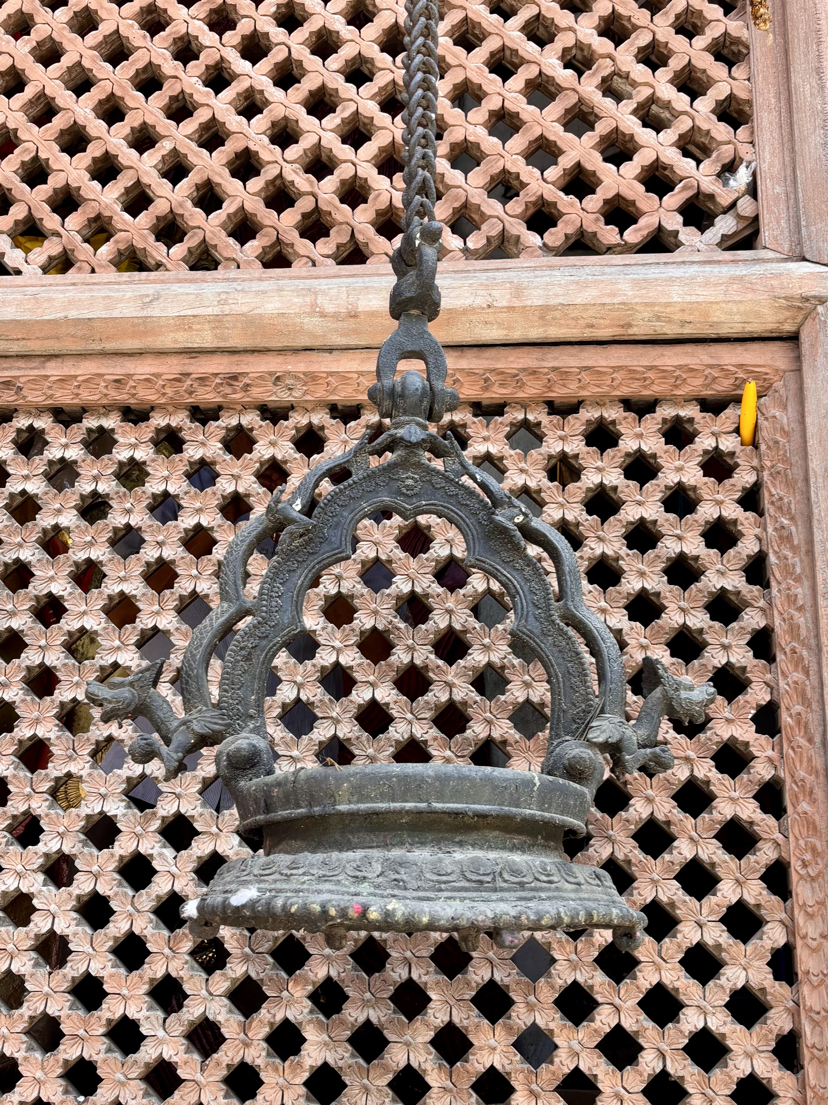 An intricately designed antique metal bell hanging in front of a beautifully carved wooden lattice window.