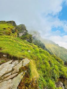 A lush green hillside rises steeply, dotted with patches of vibrant grass and small wildflowers. The terrain is uneven, with large, rugged stones protruding from the ground. 
