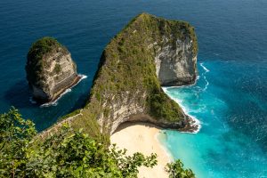 The famous T-Rex-shaped cliffs at Kelingking Beach on Nusa Penida, Indonesia. The rocky cliffs protrude into the blue sea, and a beach is visible in the bay they create.