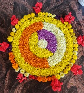 A vibrant floral rangoli on a dark floor, featuring concentric circles of yellow, orange, white, and purple flowers, with red hibiscus accents.