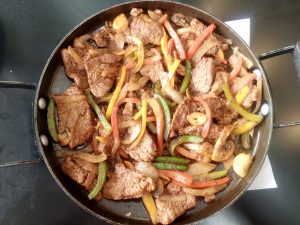Sliced beef cooked with onions, peppers, and mushrooms in a round pan.