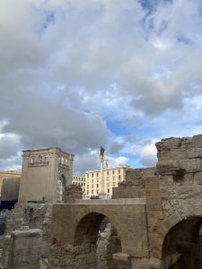 Ancient stone ruins with arches in the foreground, against a backdrop of partly cloudy skies. A modern building and column are visible in the distance.
