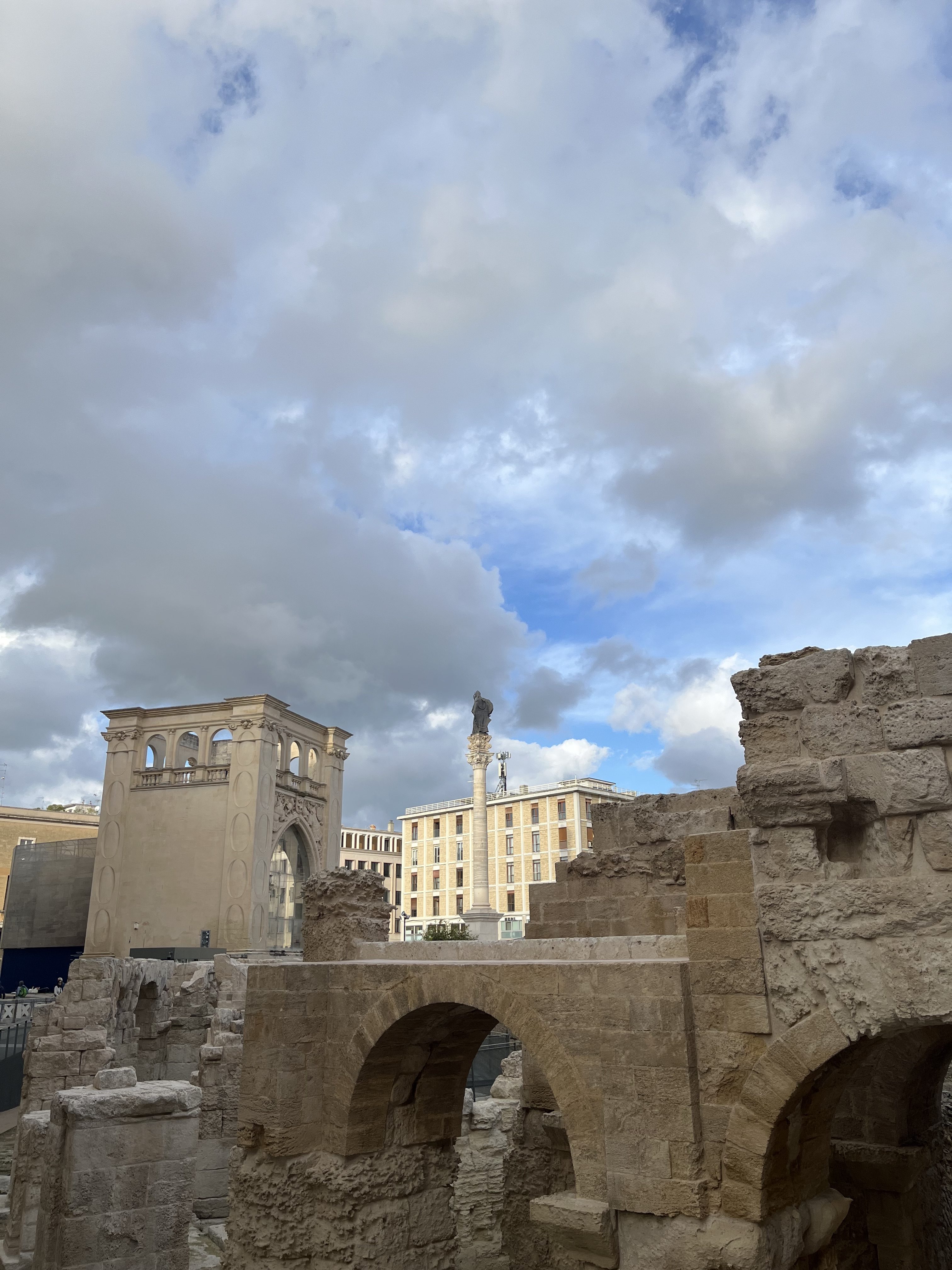 Ancient stone ruins with arches in the foreground, against a backdrop of partly cloudy skies. A modern building and column are visible in the distance.