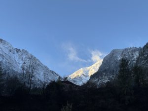 Snow-capped mountains under a clear blue sky, with sunlight illuminating the peaks. Dark silhouettes of trees and a roofline in the foreground.