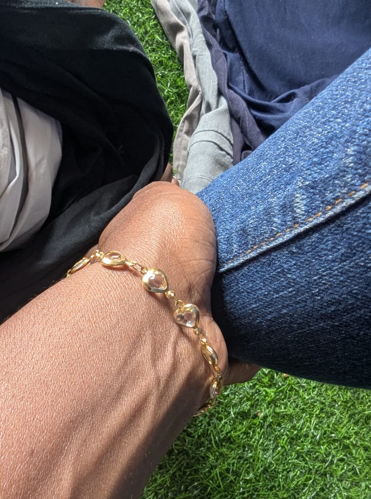 A hand wearing a gold link bracelet with heart shapes rests on blue denim-clad knees.