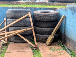 Stacked old tires against a blue wall, next to wooden planks and a leaning stone slab.