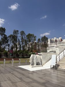 A white marble elephant sculpture decorated with ornate carved trappings stands on a square plinth at the entrance to Trimandir, Pune, India. A broad marble staircase with glass railings rises to the right, with a tree-lined lawn and residential buildings visible behind under a vivid blue sky.