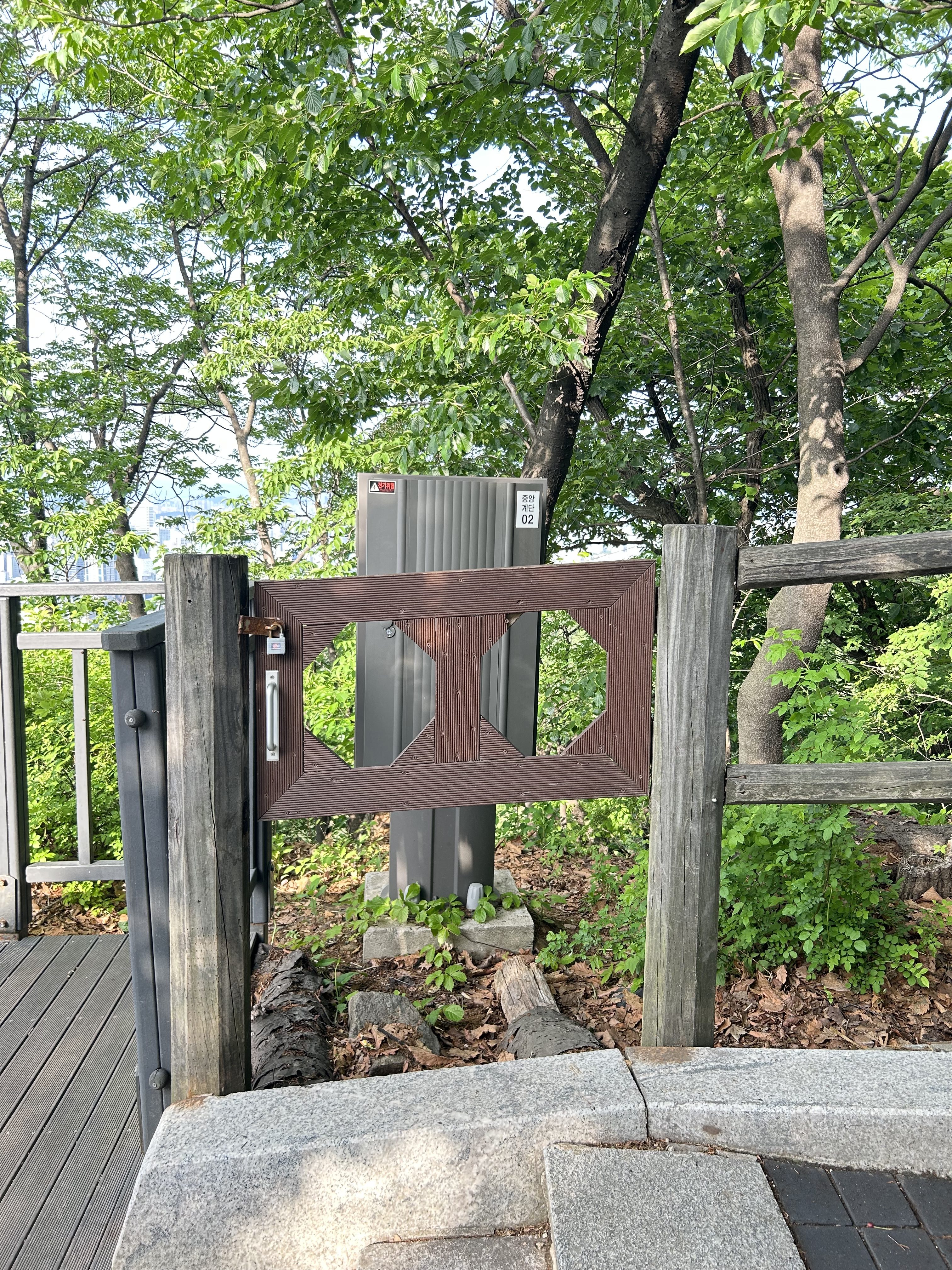 A wooden gate with a geometric design opens to a pathway surrounded by lush green trees. Behind the gate, a tall, gray utility box stands among the foliage, partially obscured by the vegetation.