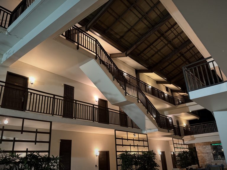 A multi-level indoor hotel atrium at night, featuring open corridors with black railings, warm wall sconce lighting, an central angled staircase, and a high, wood-paneled ceiling structure.