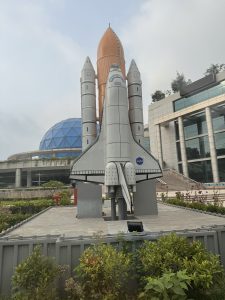 A full-scale NASA space shuttle replica with an orange external fuel tank displayed outdoors in front of a modern building with a blue geodesic dome, surrounded by green shrubs and gardens.