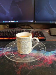 A decorative cup of milk tea on a glass saucer sits beside a red-lit keyboard and dual monitors, creating a cozy desk setup.